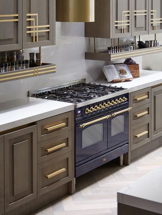 kitchen with brass detailing on cabinetry, blue range, pale wood herringbone floor, white marble countertops and backsplash, drop down shelving under wall cabinets for spices