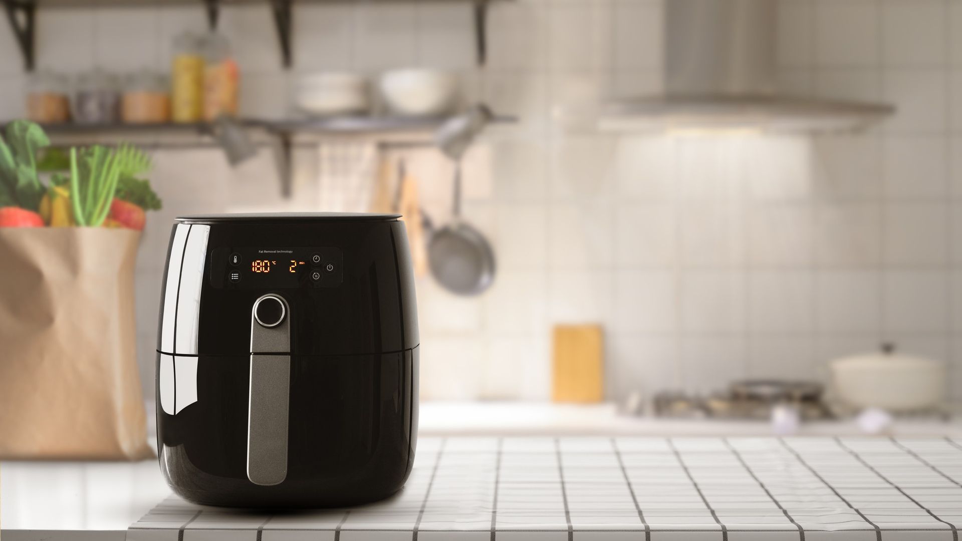 a black air fryer on a white kitchen worksurface, with a kitchen behind and utensils hanging up on a wall