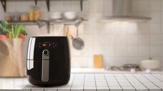 a black air fryer on a white kitchen worksurface, with a kitchen behind and utensils hanging up on a wall