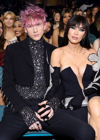 MGK wears a black suit with silver embellishments and has pink hair while Megan Fox wears a strapless black dress featuring silver flowers