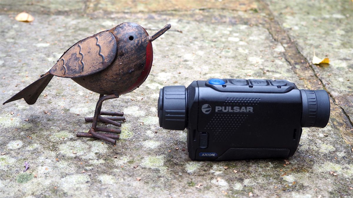 Pulsar Axion XQ30 PRO night vision monocular on a stone floor next to a toy model of a bird