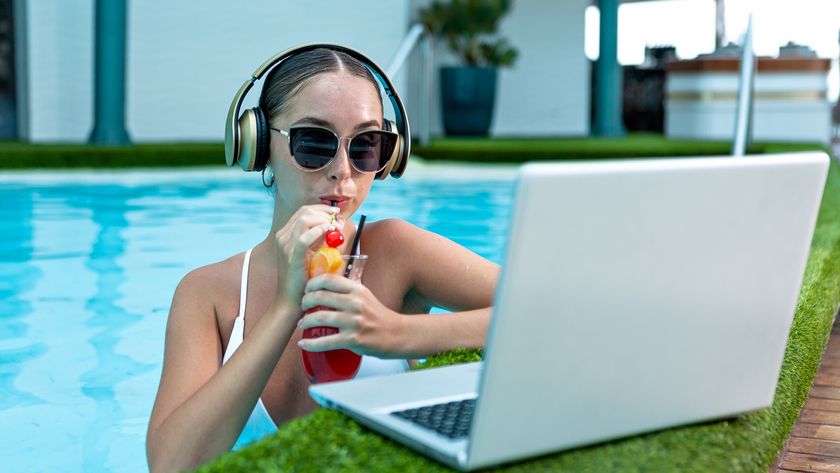 Young woman in sunglasses and wireless headphones drinking fresh cocktail and watching video on laptop while in a swimming pool.