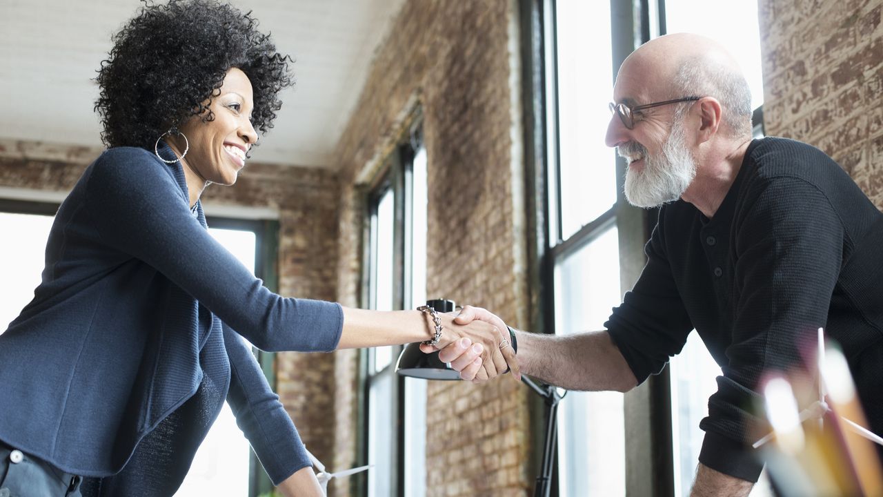 A businesswoman shakes hands with a businessman across a desk in an office.