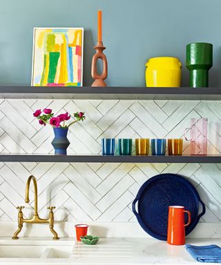 Colorful corner of a kitchen. Blue painted walls, herringbone white tile splashback, two dark wooden shelves decorated with colorful ornaments, artwork, vases and kitchenware