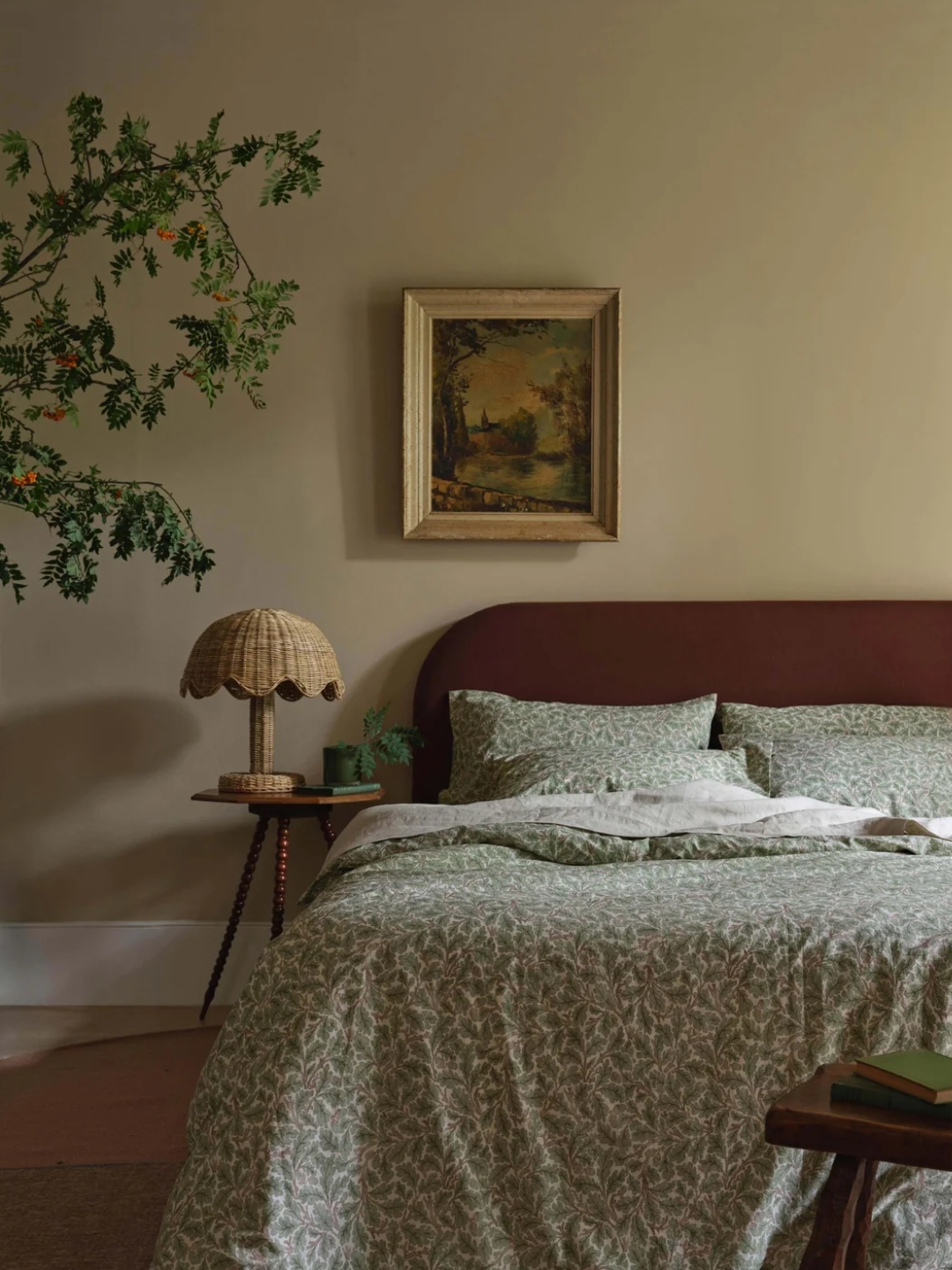 Piglet in Bed's green floral bedding on a red bedframe in a beige room. There is a rattan table lamp on the side table next to the bed, art hanging above the bed, and a tree in the corner.
