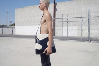 Topless man holds leather tote bag in industrial setting, part of Ferragamo ‘A New Dawn’ capsule collection
