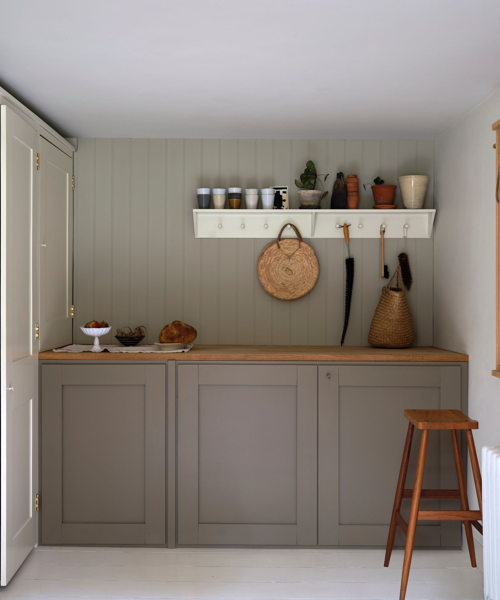 Kitchen painted in Farrow &amp;amp; Ball's Mouse's Back, wooden stool, wooden countertop, a white shelf holding ceramics, and small baskets hanging up.
