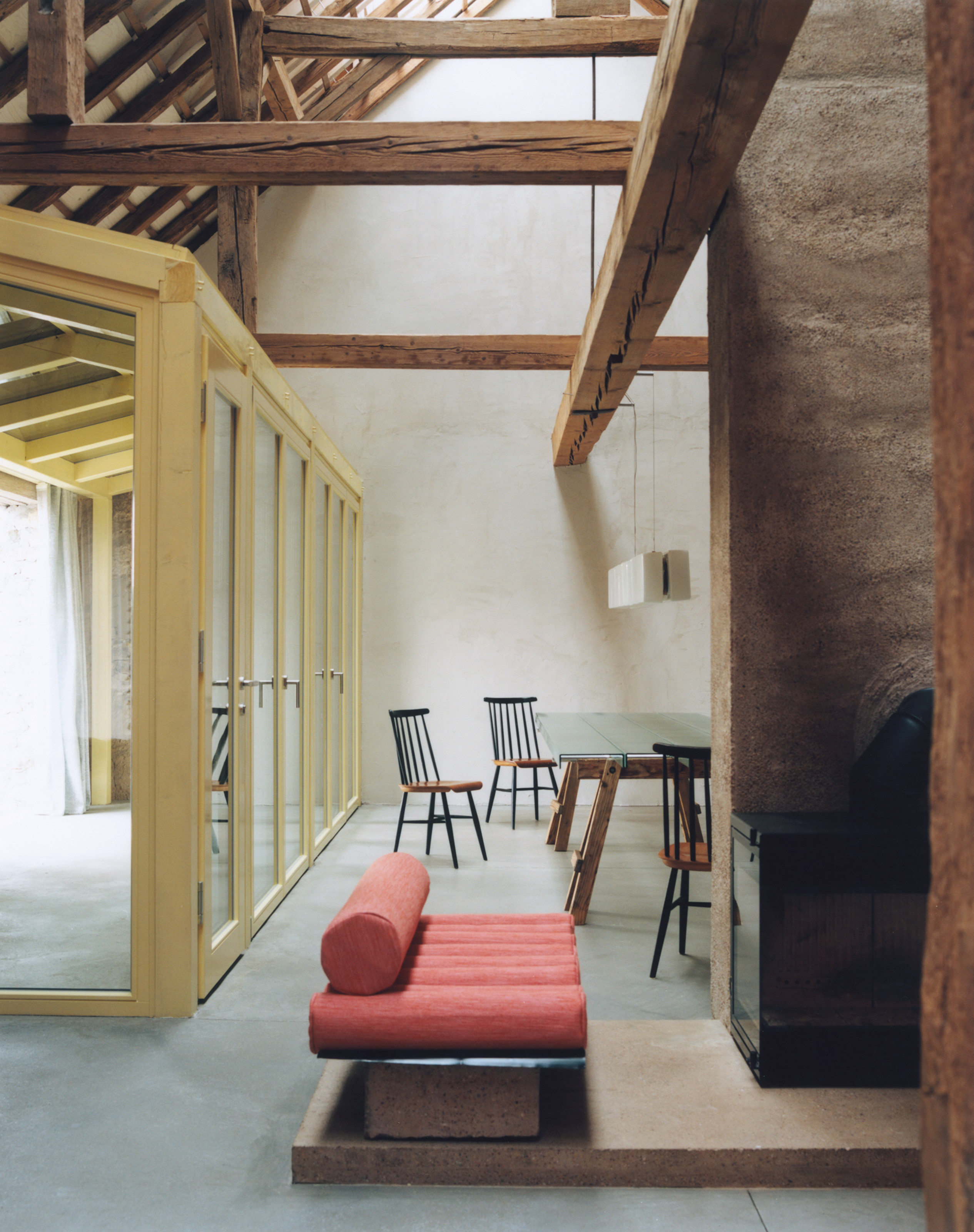 Open-plan living-dining area with yellow-framed internal glass walkway that leads through to a courtyard with beams overhead, karge concrete chimney in the center and dining table in the background with a red modern bench in the foreground