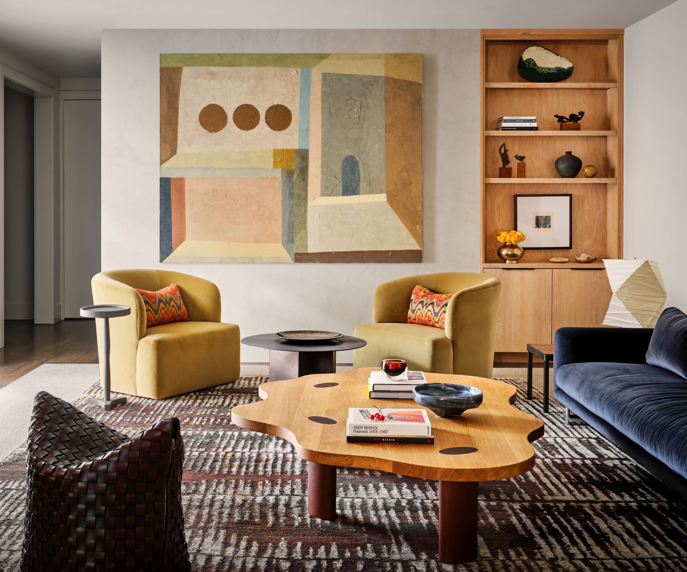 Living room with wood book case, yellow velvet amrchairs, blue velvet sofa, patterned rug, wood coffee table and woven pouf
