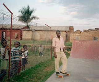 A man standing next to a skate board on a skate board ramp with kids standing by a fence watching.