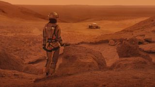 a person in a spacesuit on a red, dusty planet looks at a wheeled vehicle in the distance