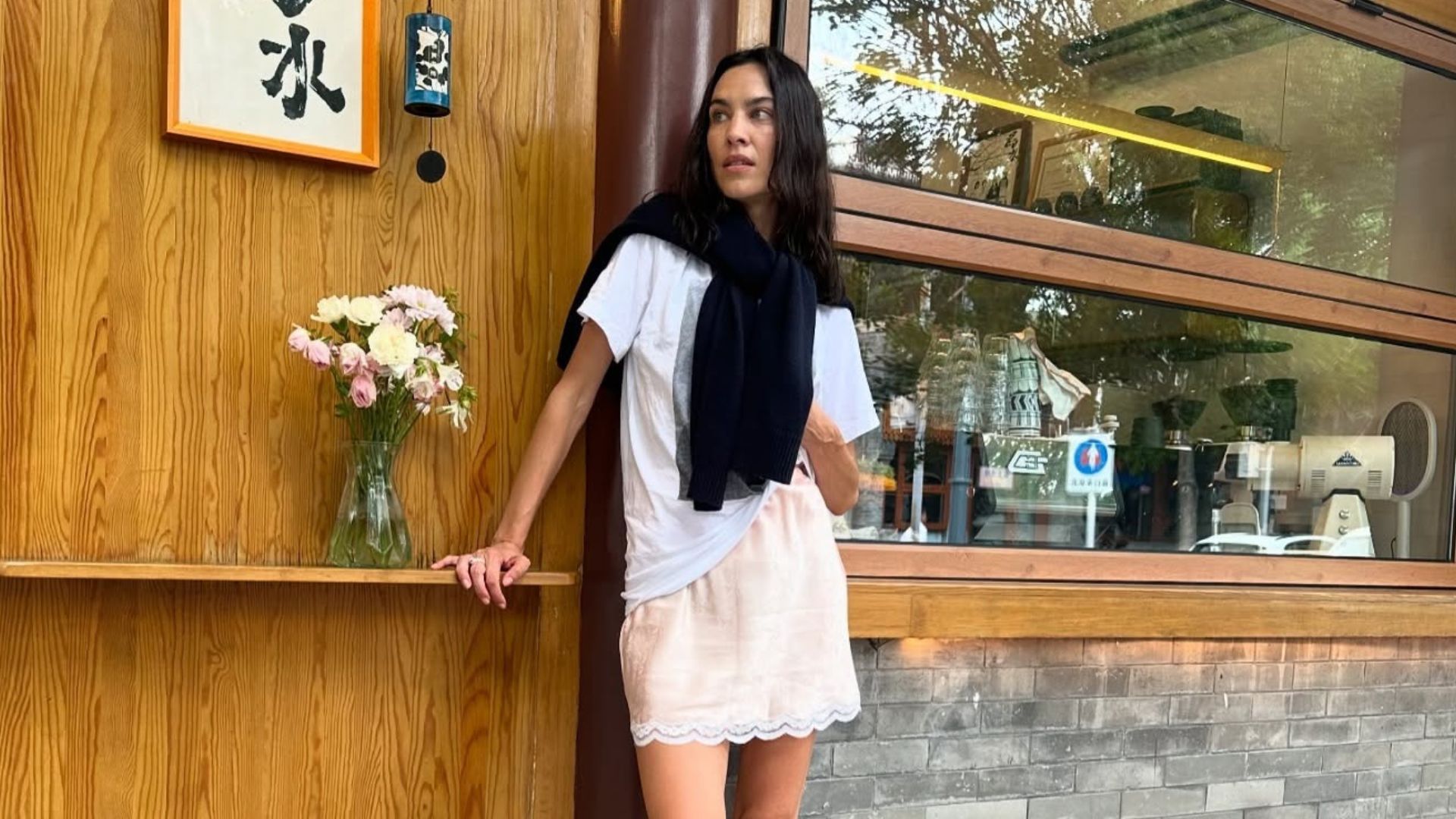 Alexa Chung stands outside wearing a pink satin miniskirt with lace-trim detailing with black trainers, a white t-shirt and a jumper wrapped around her shoulders.