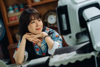 a woman rest her head on her hands and smiles in a still from the k-drama when the camellia blooms