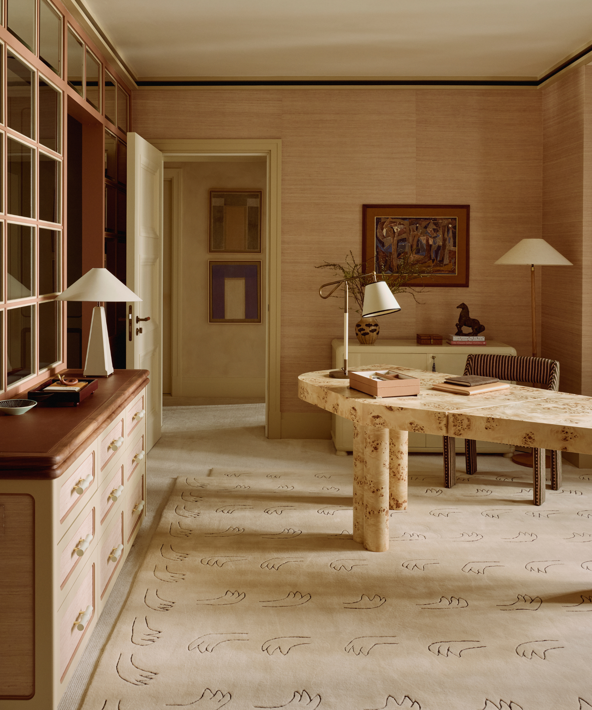 Neutral office space featuring an asymmetrical burl wood desk, linen-shaded table lamps, wall art, textured wallpaper, a figural motif beige rug, and a striped office chair