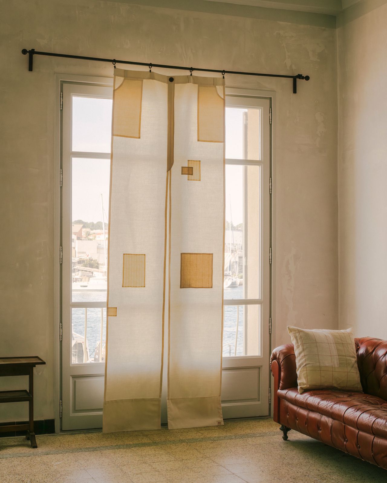 A bright but older living room with French doors that have a noren-inspired linen curtain hanging in front of them that is shaped like a pair of pants.