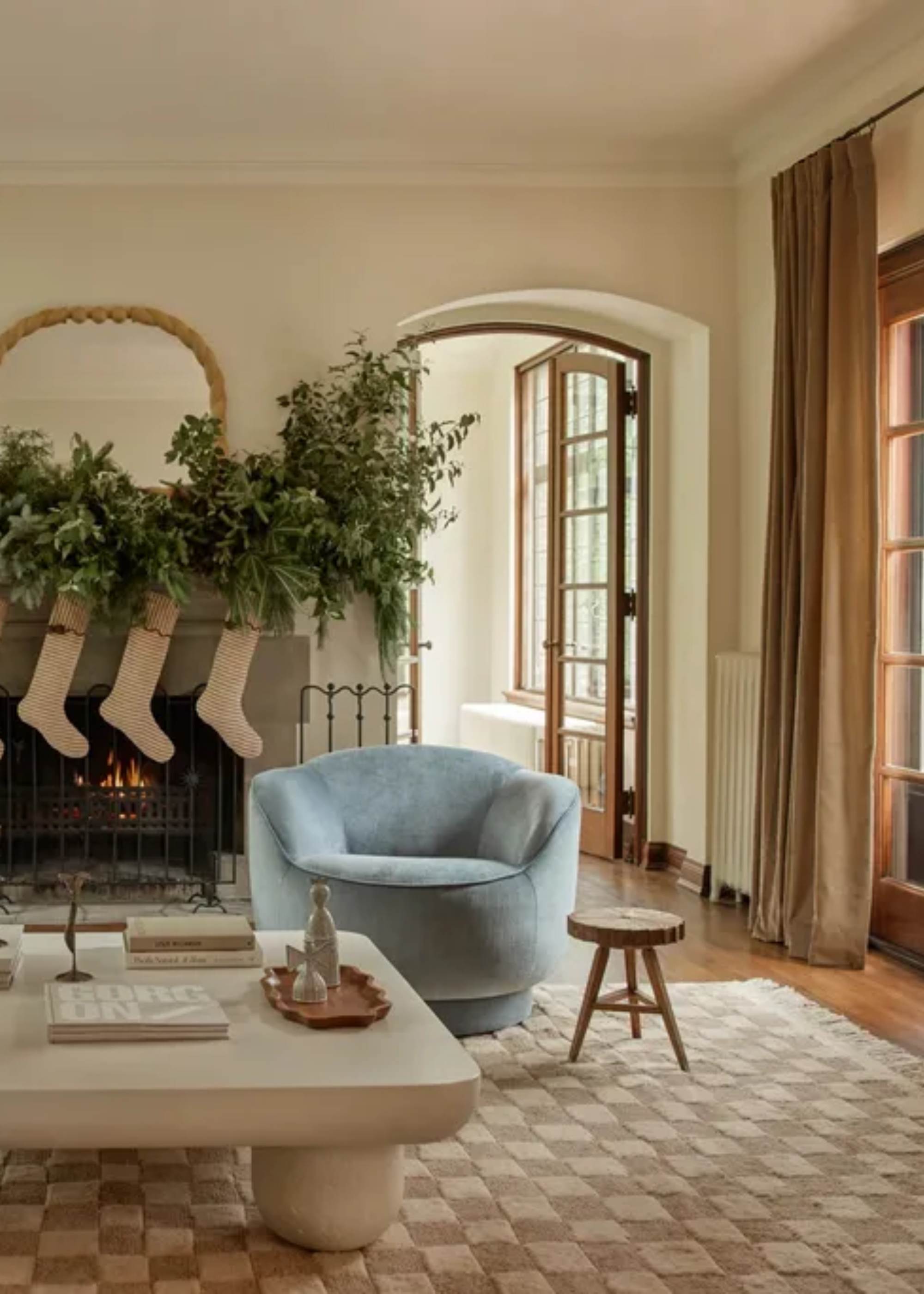 A living room with a checkered rug, a pale blue sofa chair, a wooden stool, a low white stone coffee table and a fireplace with a row of stockings and garland