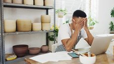 A business owner sits in front of his laptop at his desk with his face in his hands.