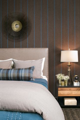 Bedroom with bed with upholstered headboard, mirror above on wall with brown and blue stripe and nightstand