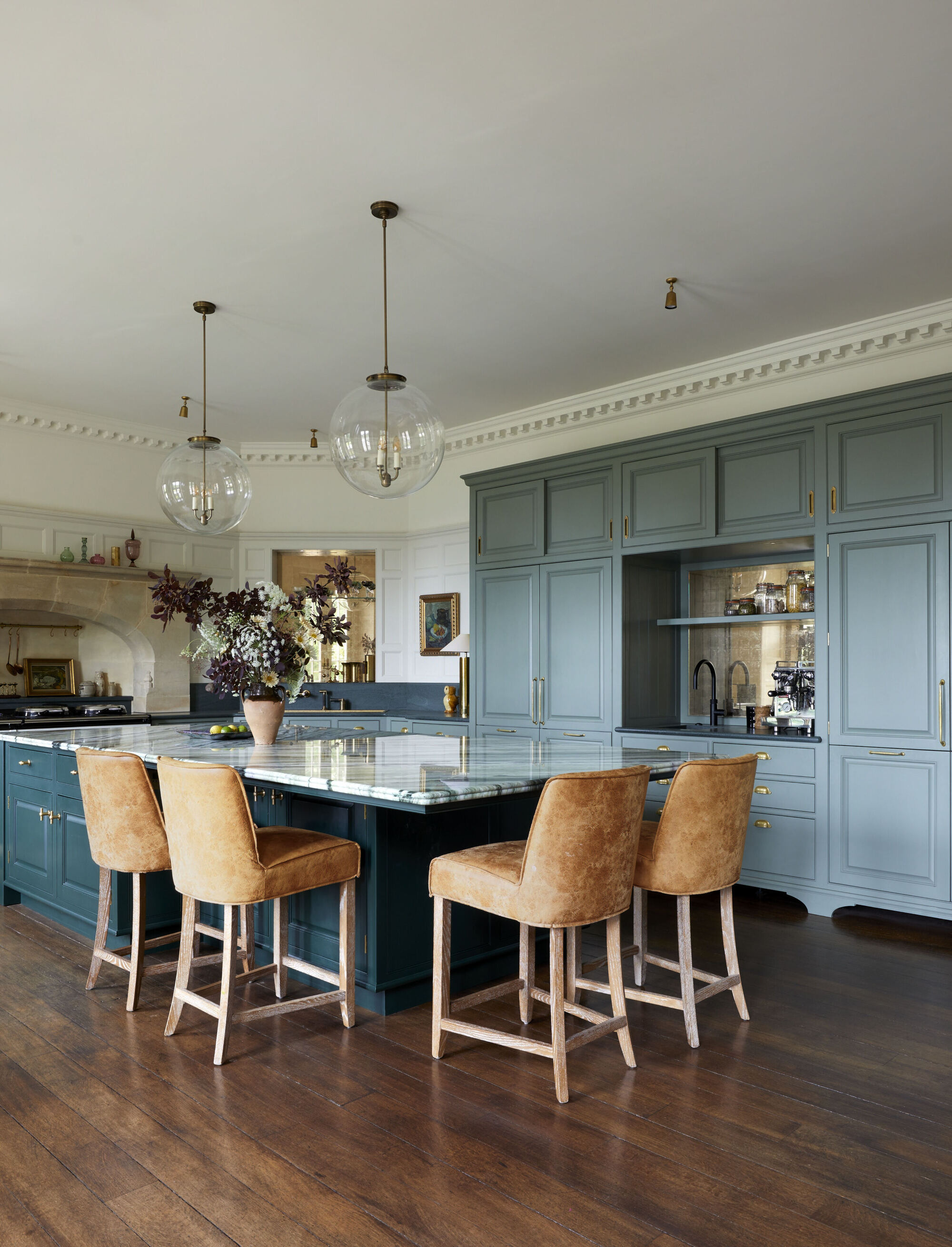 green kitchen with traditional shaker style cabinetry, dark timber floors, crown molding, marble island bench with tan bar stools, pendant lights hanging over bench, and vase with flowers