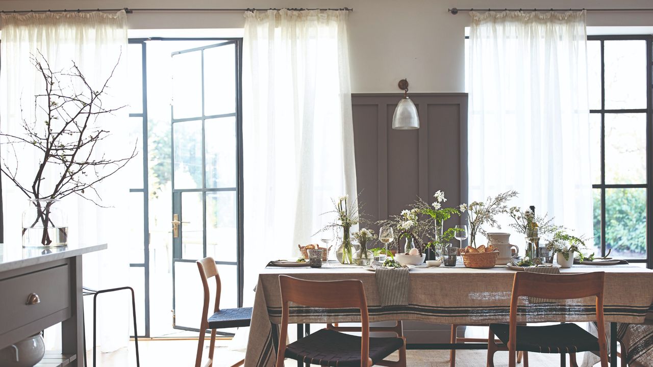 A dining room with a set table and two large windows covered by sheer curtains