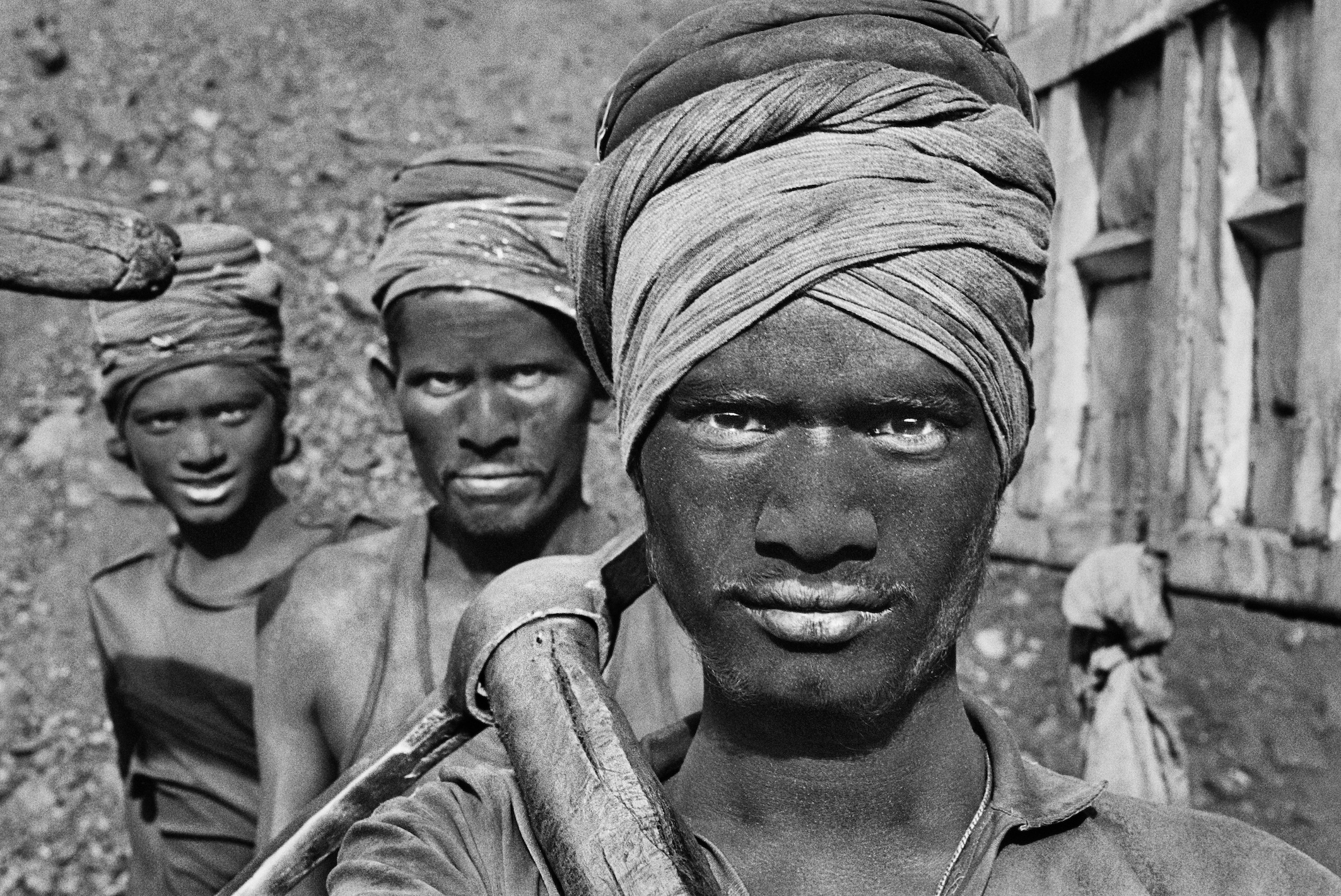 A close-up, black-and-white photograph shows three coal miners with dark, coal-dust covered faces and head wraps, with the man in the foreground looking directly at the camera while holding a shovel over his shoulder.