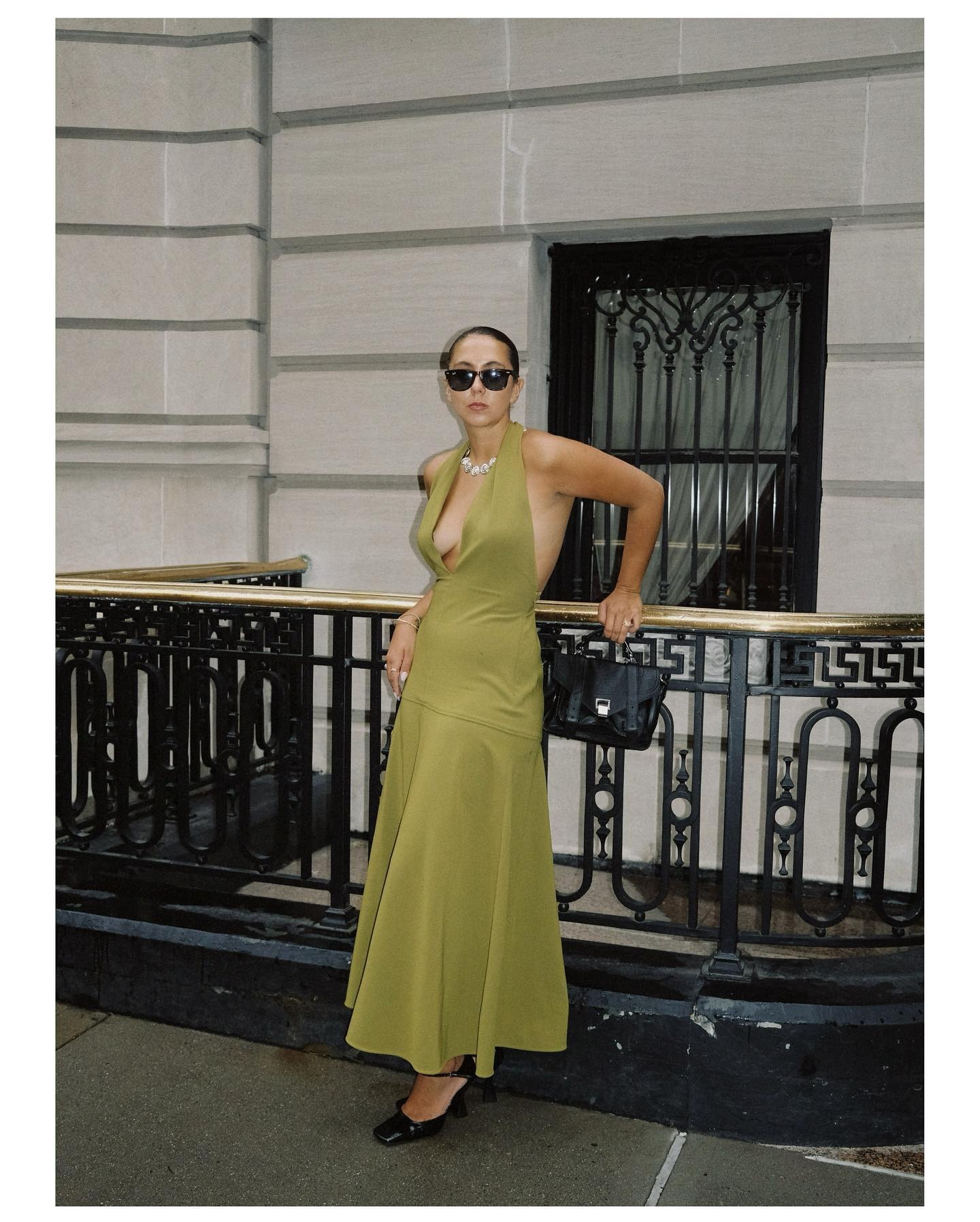 woman wears green halterneck maxi dress with black accessories leaning against a railing.