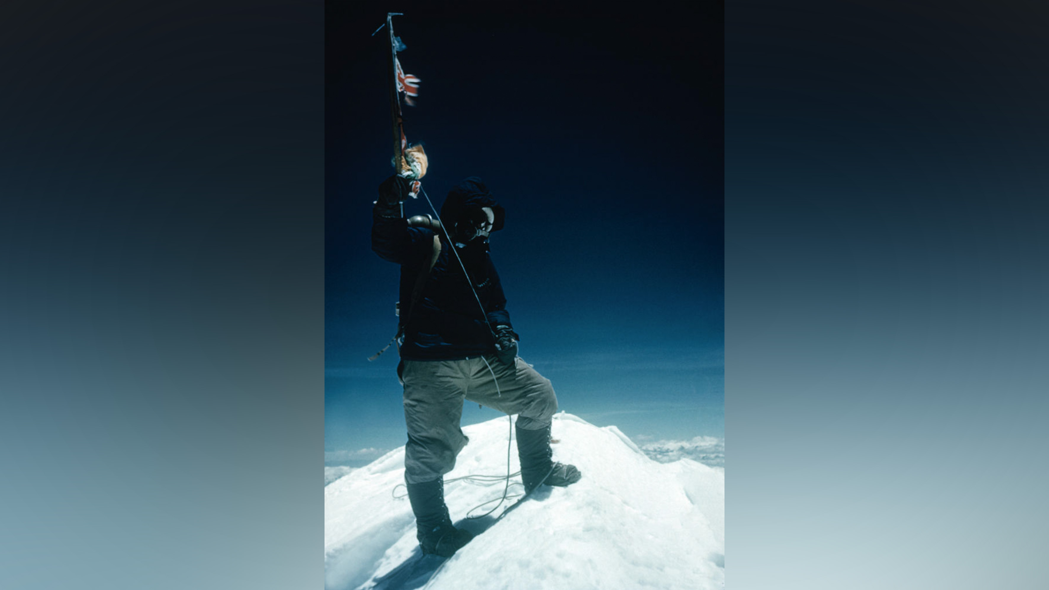 Man in extreme-cold clothing stands atop mountain holding ice axe in the air.