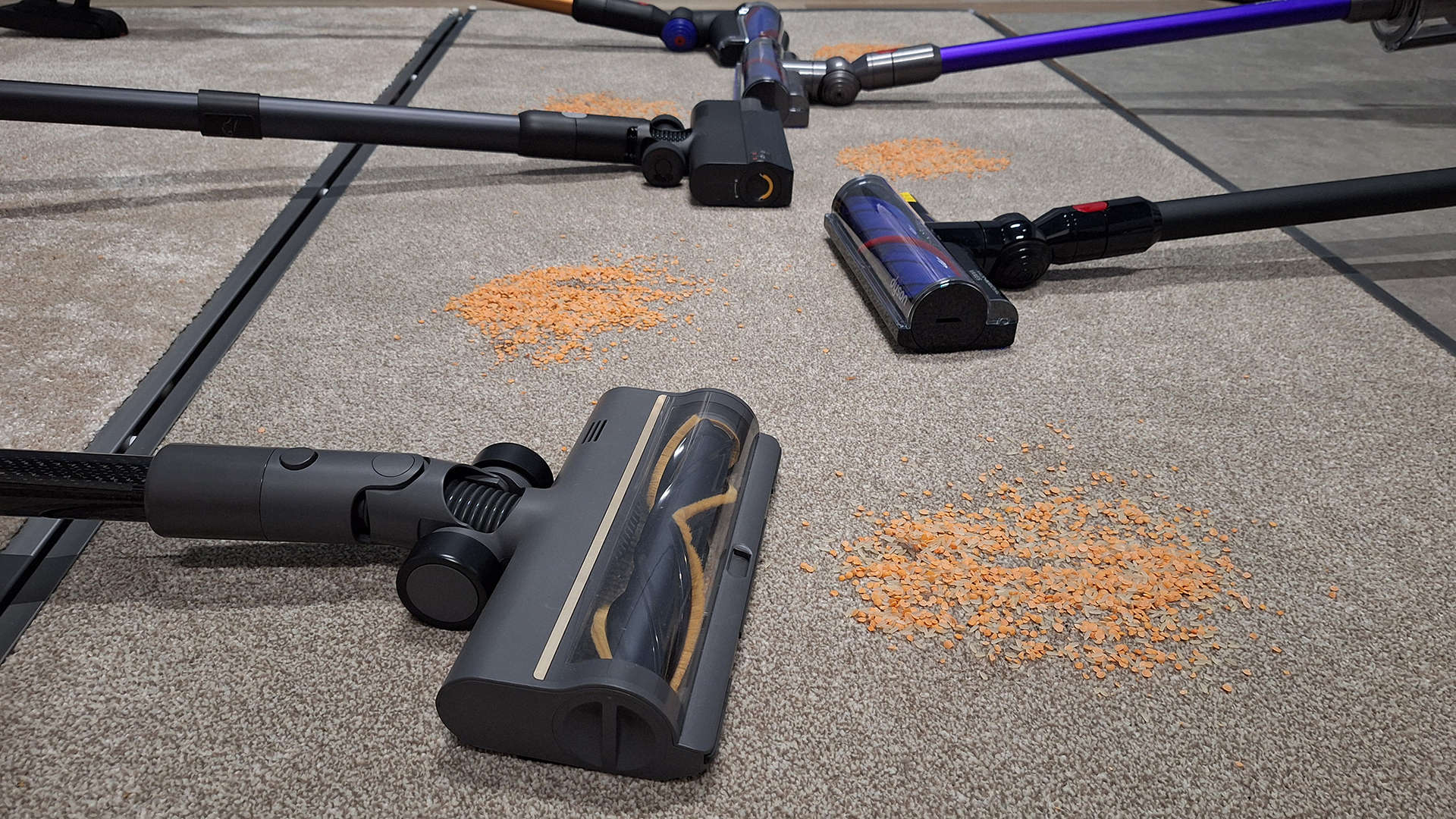 Various cordless stick vacuums lined up on a carpeted panel, with dry lentils and rice spilled in front of each floorhead