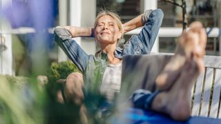 Woman happily sitting in the sunshine in back garden with feet up on wooden bench and book in her lap