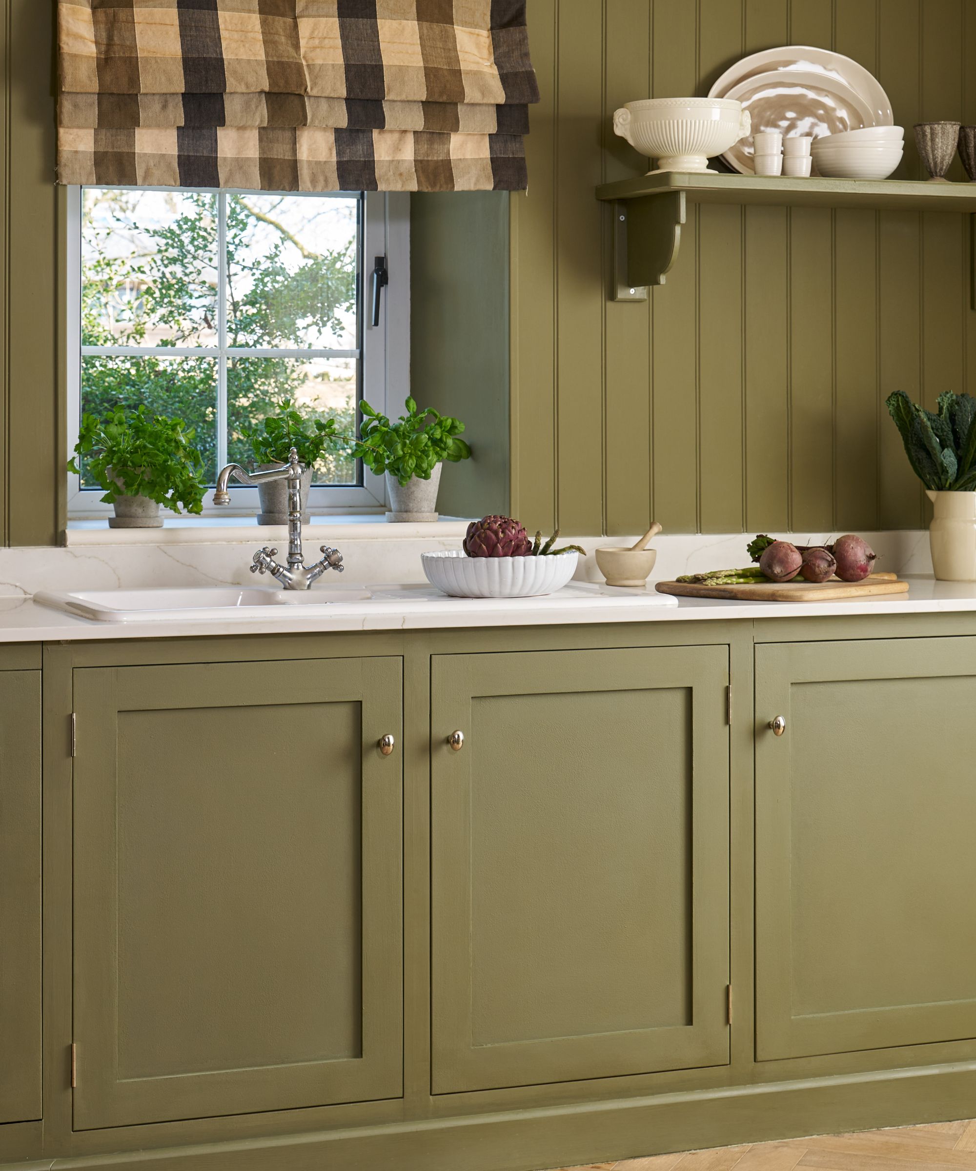Olive green shaker kitchen with panelled walls and matching cabinetry. A white worktop sits beneath a window with a checked Roman blind, surrounded by potted herbs, vegetables and simple crockery, creating a warm country-style atmosphere.