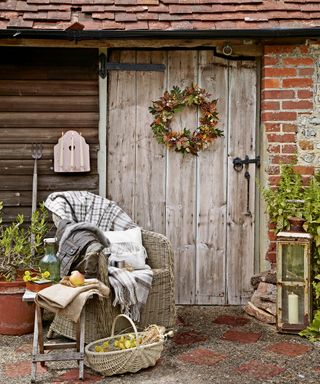 A rustic front porch area with a wooden door, a rattan armchair with a blanket, and a basket of vegetables
