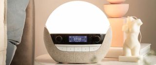 The Lumie Bodyclock Luxe 700FM sunrise alarm on a warmly-lit bedside table. 