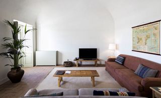 Private living space in the São Lourenço do Barrocal hotel. Burnt brown couch to the right, with a map on the wall above it. A wooden coffee table in the center, with a beige rug on the floor. A big plant to the right, and a tv to the farthest wall.