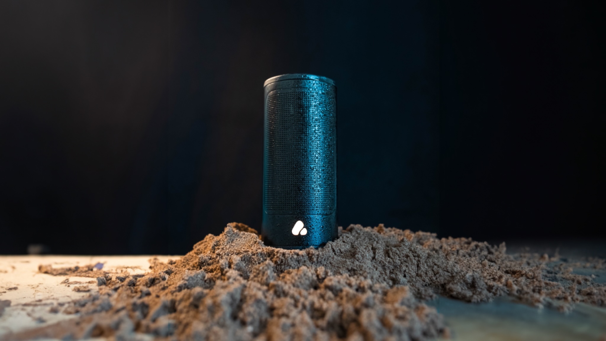 A Pine Beat speaker in black, on a pile of sand, in front of a black background