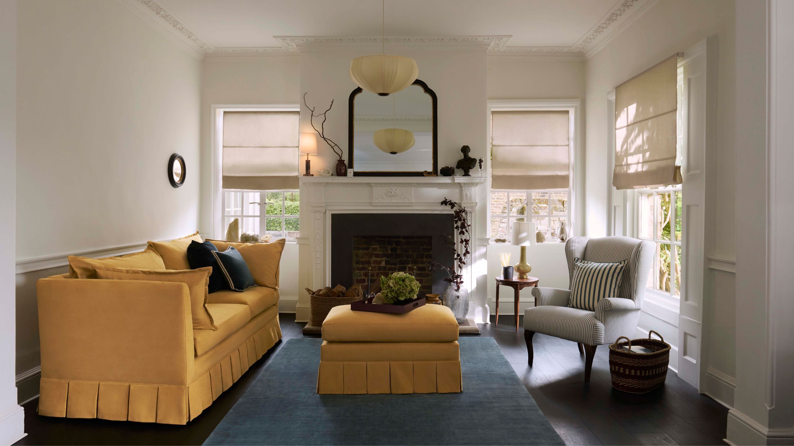 living room with yellow pleated sofa and ottoman, blue rug, fireplace, armchair with striped cushion, arched mirror and roman blinds over windows