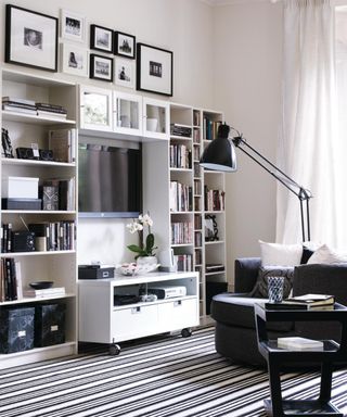 A black and white small theatre room with white tall bookcases and shelves, black and white wall art prints, a TV, TV stand with wheels and decor, and a black floor lamp, round armchair, and side table with books on