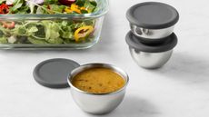 Three stainless steel condiment containers with grey silicone lids, on a white marble counter beside a glass dish of salad. 