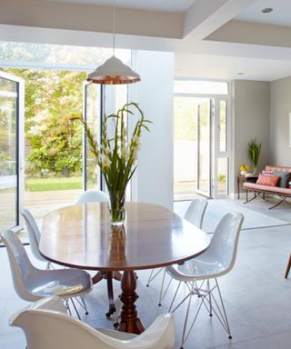 A white dining room with an door opening to a garden, a white and rose gold pendant light hanging over a curved wooden dining table with white plastic chairs underneath it and tall flower stems on top of it