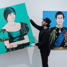 A man wearing black hangs a turquoise pop art Andy Warhol painting of Princess Diana next to one of Prince Charles