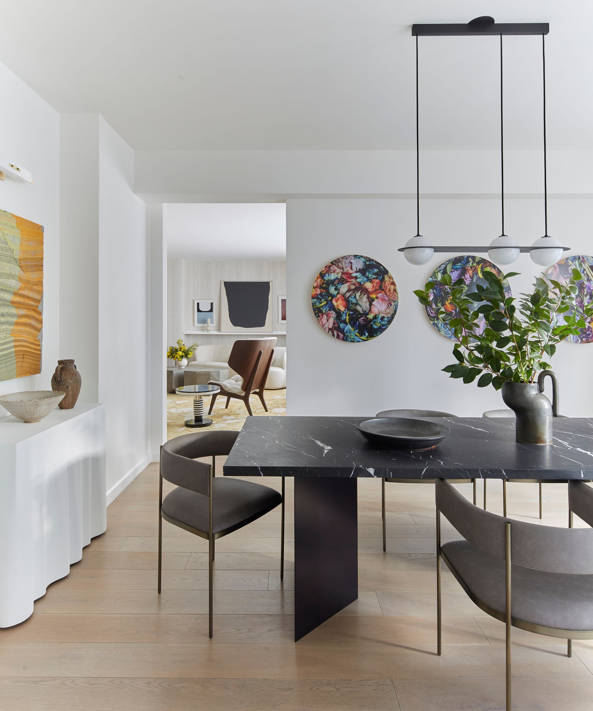 Minimalist dining space with black marble table, curvaceous dining chairs, geometric pendant hanging over table, artwork on wall in background, view into living room