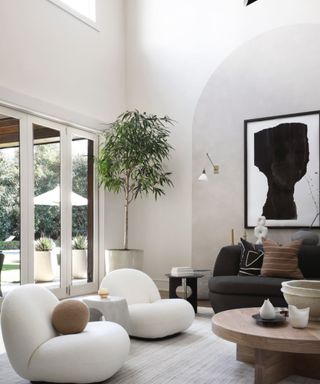 A white living room with two white sculptural chairs, a tree in the corner, and sliding glass doors leading outside