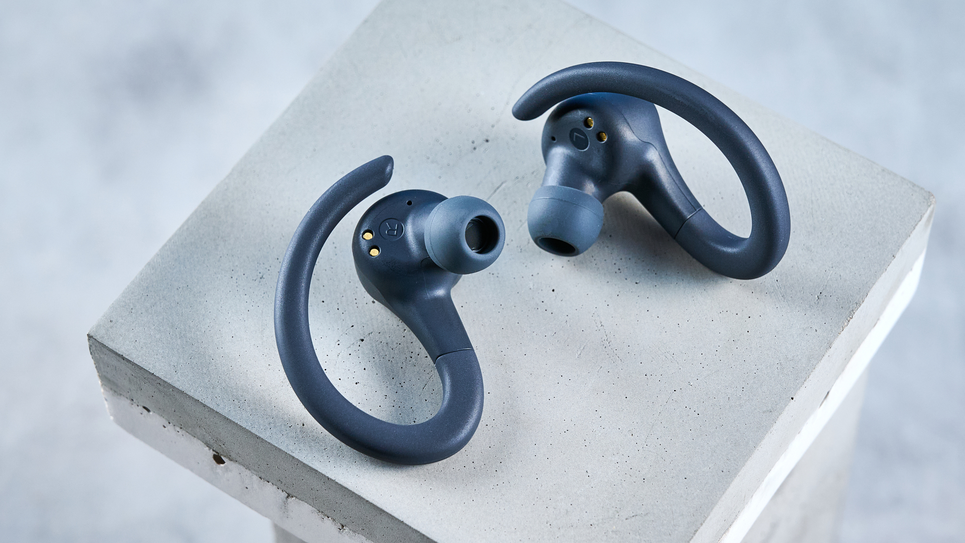 the jlab go sport+ earbuds photographed against the blue tom's guide background, showing charging case and wearing style