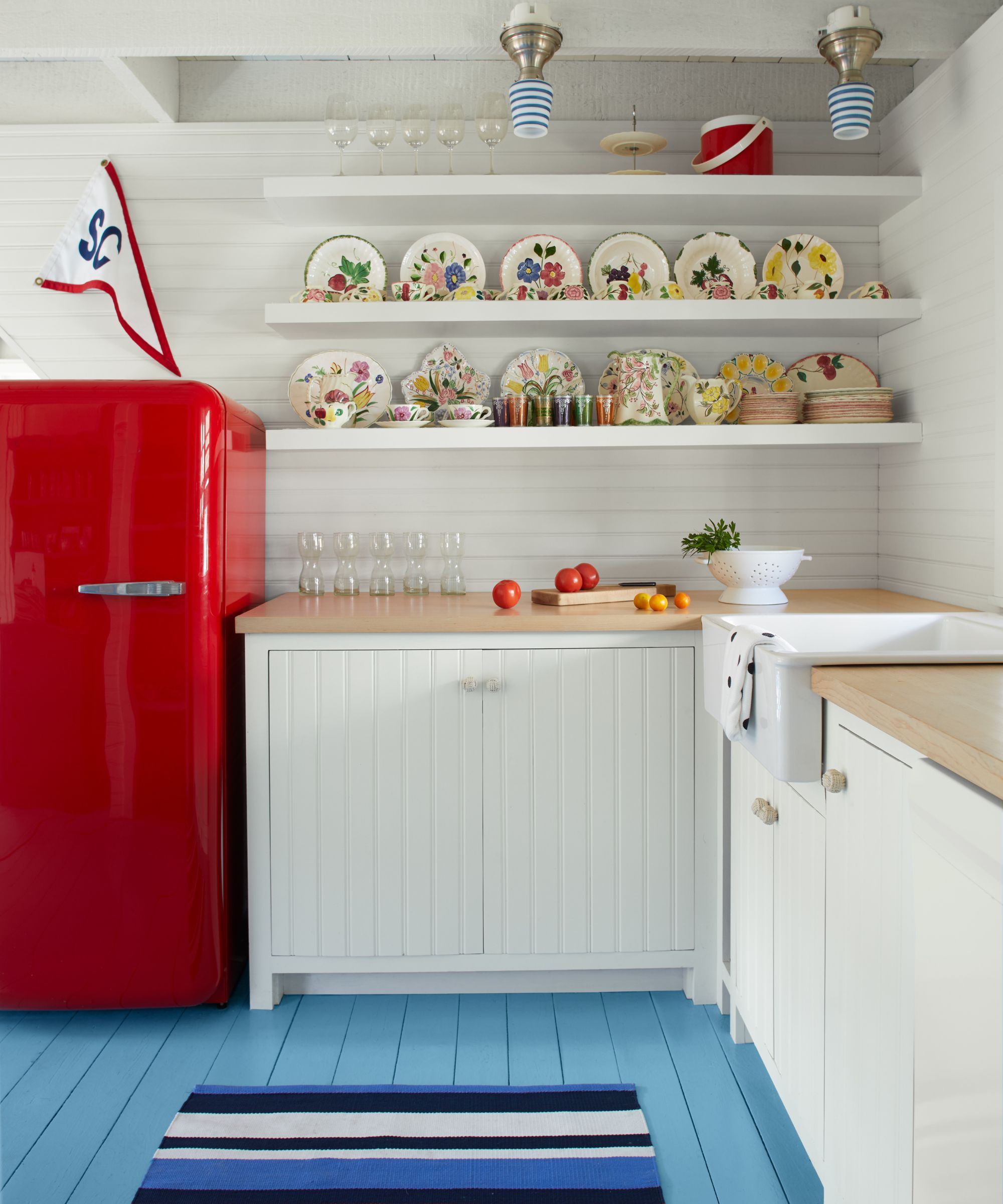 A small coastal kitchen with white cabinets and walls, a blue painted floor, and a red fridge