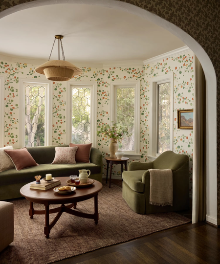 a room with vine-like wallpaper, a green accent chair and sofa, a wooden coffee table, and a burgundy rug