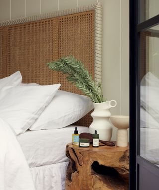 The corner of a bed, laid with white sheets; to the right, a wooden nightstand, topped with vases and fragrance bottles.