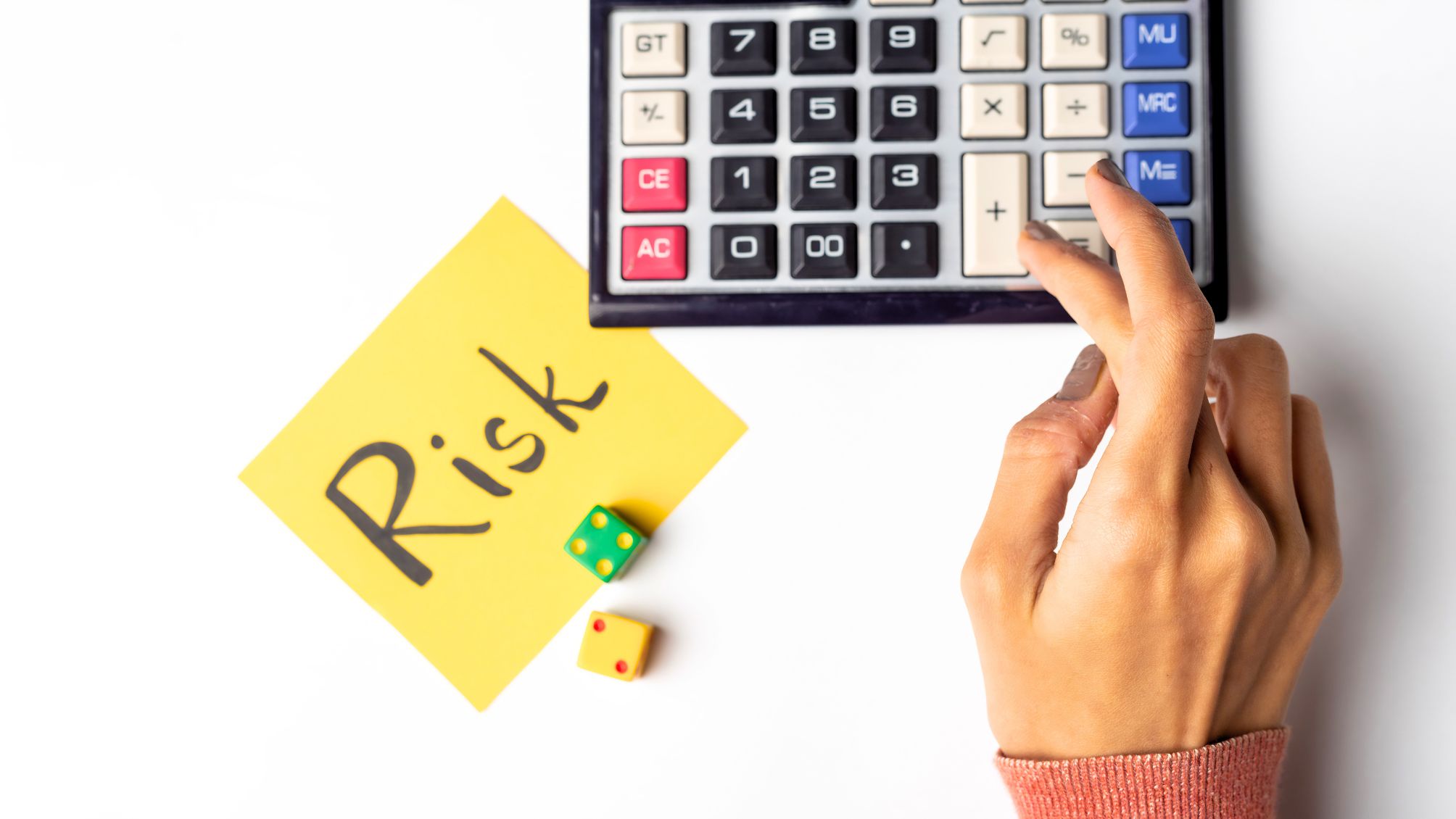 A hand using a calculator next to a sticky note and dice. The note has "Risk" written on it.