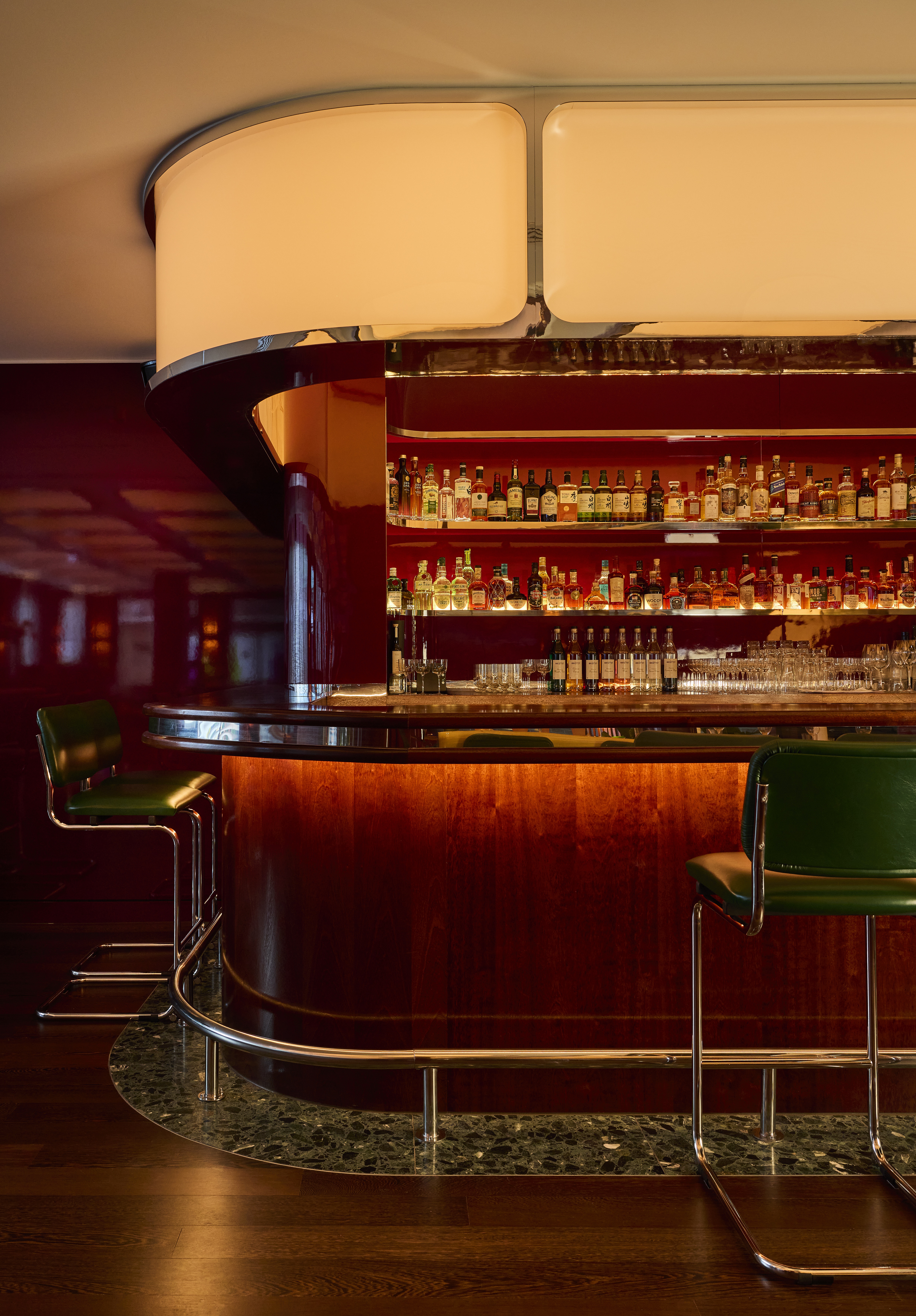 A restaurant bar features a back-lit, buttery-tinted frieze framed by sleek chrome detailing, sinuous soft lines, a 1970s palette of earthy red and green tones, and tall leathery stools.