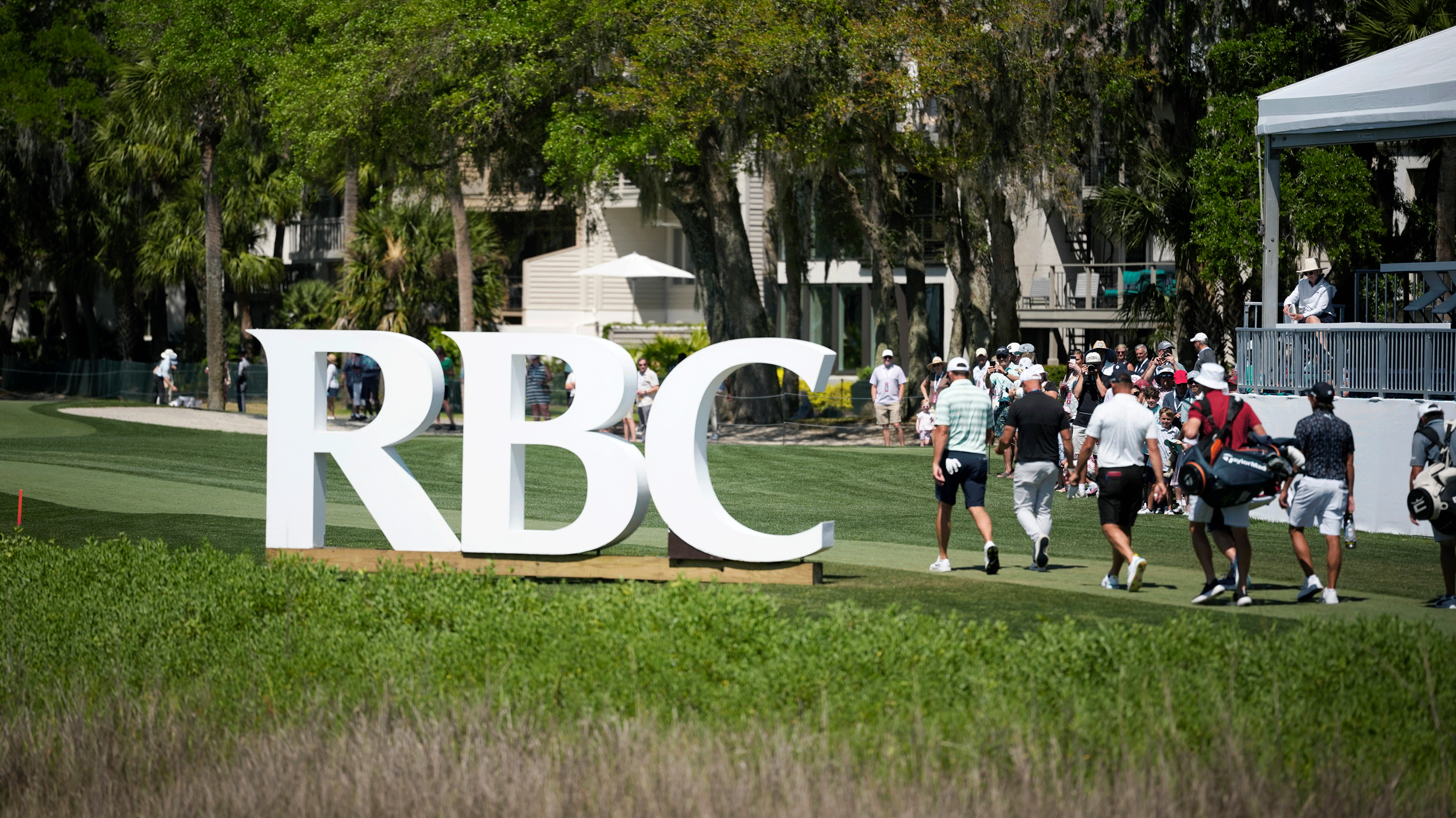 A general view of the RBC sign at Harbour Town Golf Links with three PGA Tour pros and their caddies walking down the 18th hole
