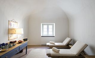 A relaxing room in the São Lourenço do Barrocal hotel. White walls with a domed ceiling. To the right, we have two beach-style beds with towels on them, to the left we have a table with two drawers in navy blue.