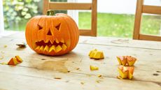 A pumpkin with a carved spooky face on a wooden table outside. Pumpkin chunks and stains on the table.